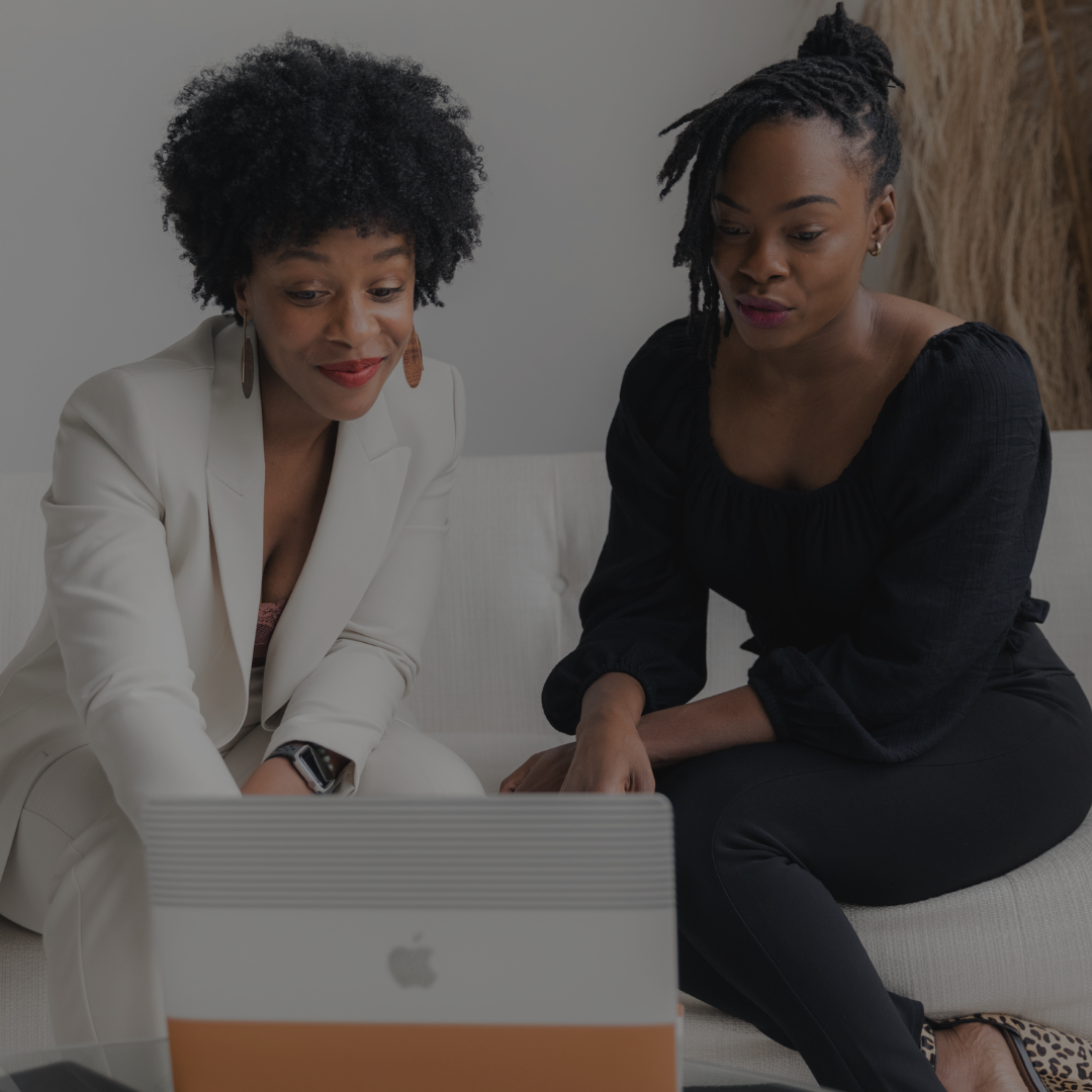 Two black women one with curly hair in a white suit helping another black woman with locs in an updo looking at a computer together on a white couch